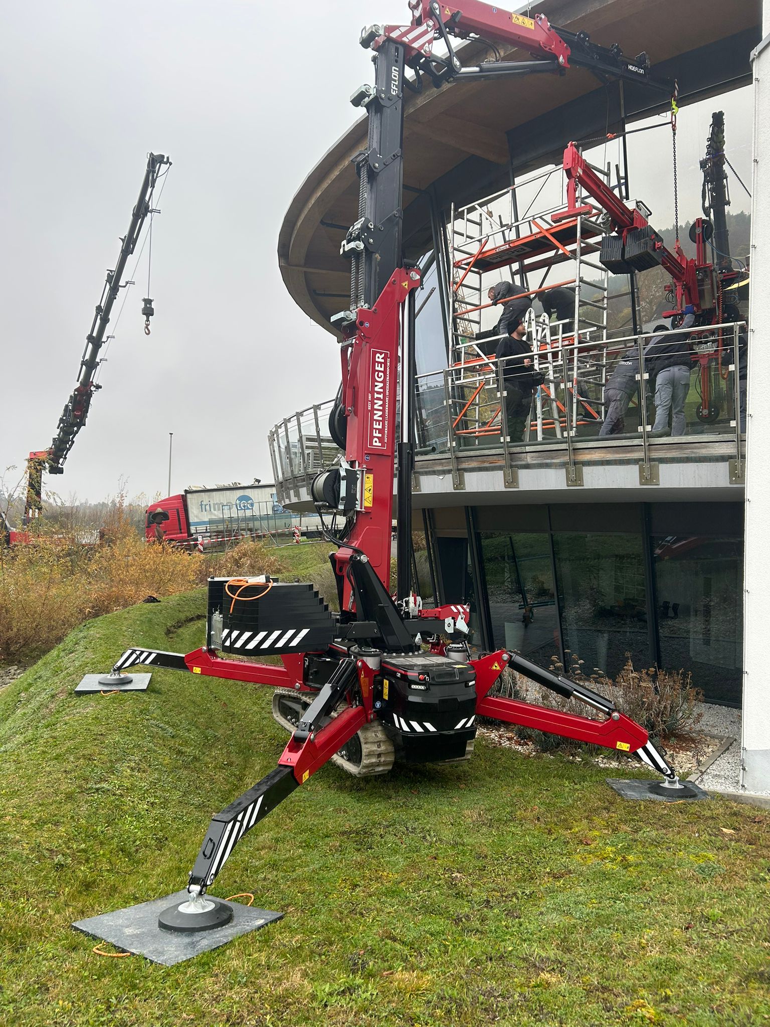 Minikran arbeitet auf Wiese bei Gebäudefassade, Ausleger vollständig ausgefahren.